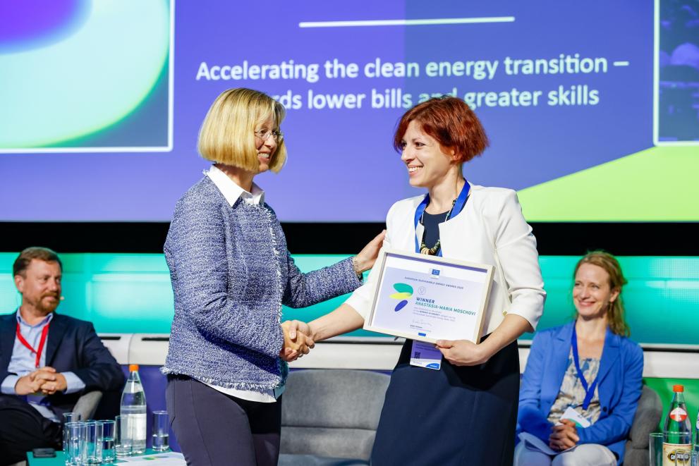 Two women shake hands at a conference, one of them holds an award. 