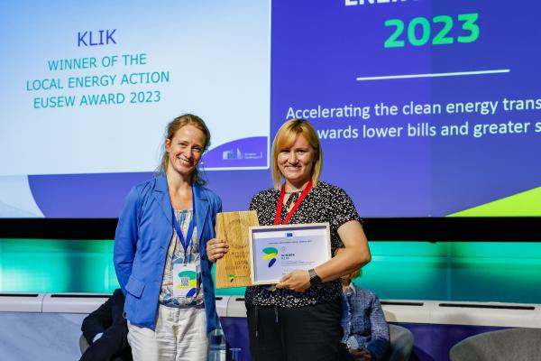 Two women standing on stage at a conference, both smiling as one of them holds an award.